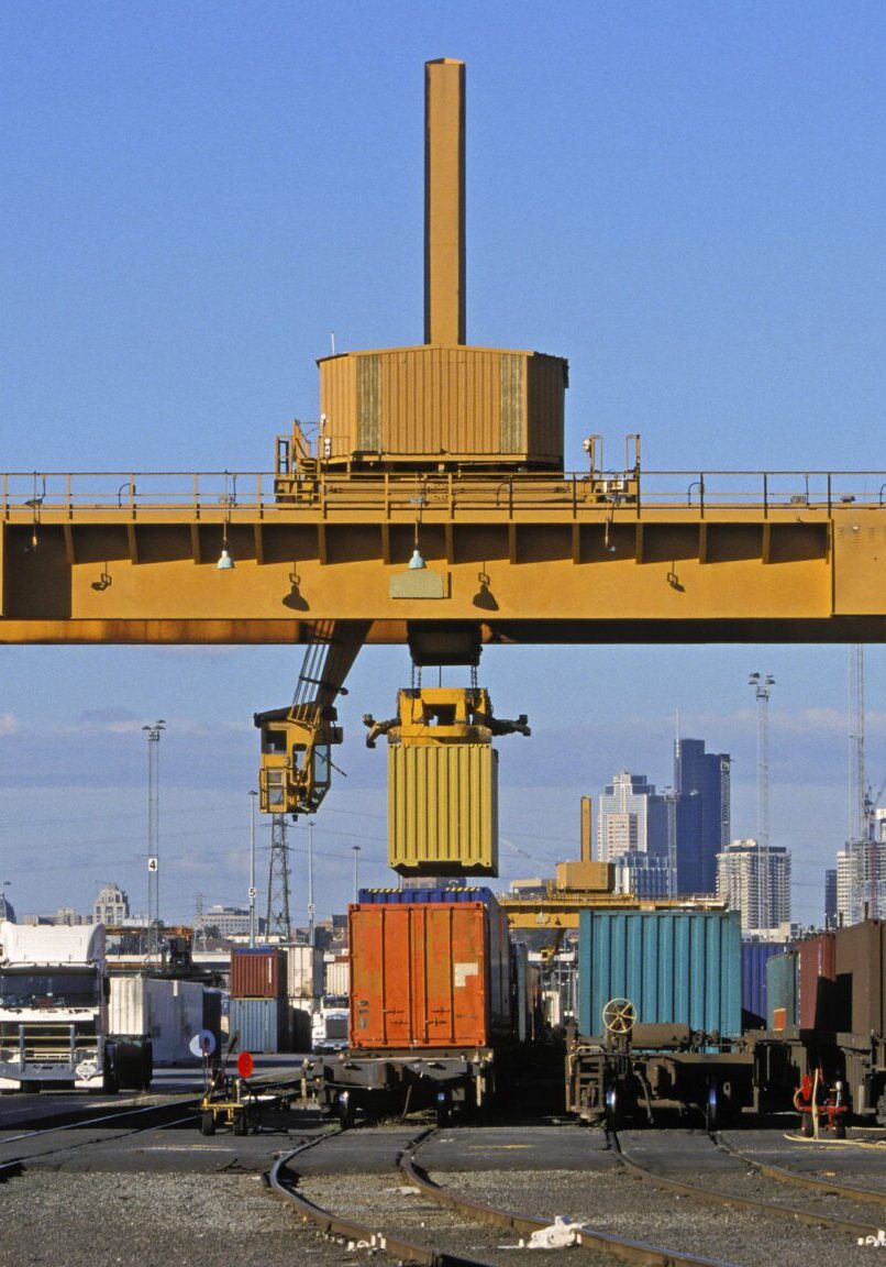 A large straddle crane loads freight containers from trucks to trains at a city intermodal hub.  High-rise buildings loom in the background.  A yellow container is in transit.  Three lines of rail wagons are being loaded with containers aa a fourth line is empty.  Trucks and stacks of containers complete the picture. Copyspace.